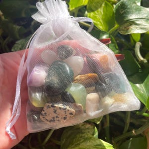 Puede incluir: Una bolsa de organza blanca llena de piedras pulidas de varios colores, incluyendo piedras verdes, marrones, negras, blancas y rosas.