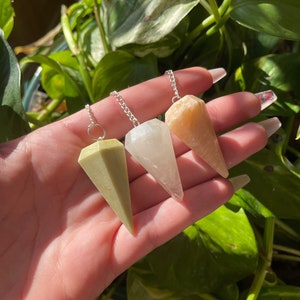 May include: Three crystal pendulums in shades of green, clear, and peach. Each pendulum is cone-shaped and suspended from a silver chain. The pendulums are held against a backdrop of green foliage, with sunlight illuminating the crystals.