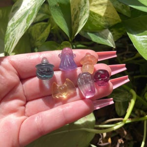 May include: A collection of small, colorful crystal figurines in various shapes, including a ghost, a bat, a skull, and a cauldron. The figurines are in shades of purple, orange, yellow, and clear, with a glossy finish. The background is a blur of green foliage.