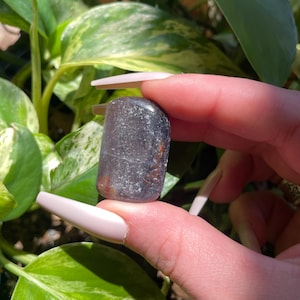 Pode incluir: Uma pedra polida, retangular, com uma cor de base cinza escuro e inclusões castanho-avermelhadas. A pedra é segurada contra um fundo de folhagem verde e amarela. A superfície da pedra parece lisa e ligeiramente refletora.
