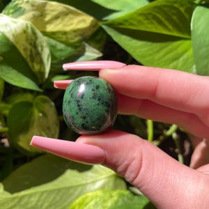 May include: A polished, spherical gemstone, primarily green with dark speckles and red inclusions. The stone is held against a backdrop of green foliage, highlighting its natural colors and textures. This gemstone is a decorative item.