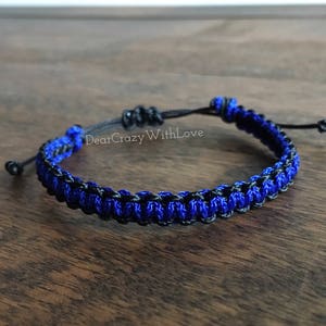 Puede incluir: Un brazalete trenzado azul y negro con cierre de nudo. El brazalete est&aacute; hecho de dos hebras de cord&oacute;n, una azul y una negra, que se tejen juntas para crear un patr&oacute;n &uacute;nico.