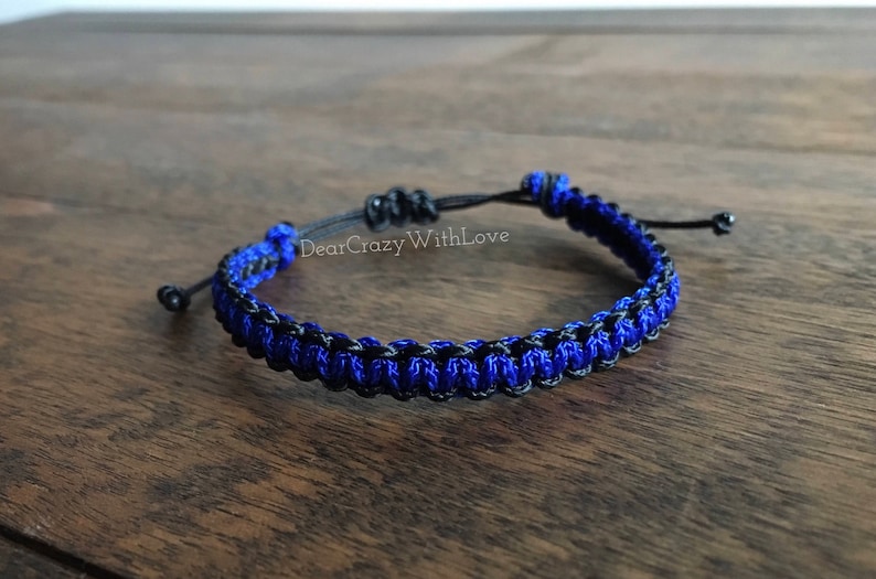 Puede incluir: Un brazalete trenzado azul y negro con cierre de nudo. El brazalete est&aacute; hecho de dos hebras de cord&oacute;n, una azul y una negra, que se tejen juntas para crear un patr&oacute;n &uacute;nico.