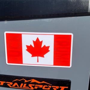 Puede incluir: Una pegatina de bandera canadiense roja y blanca en un coche gris. La pegatina es rectangular y tiene un fondo blanco con una hoja de arce roja en el centro. La bandera está rodeada por un fino borde blanco.