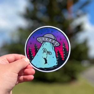 May include: A round sticker with a colourful gradient background of pink, purple, and blue. A UFO is flying over a forest with a person holding a bicycle in the air. The sticker has a white border.