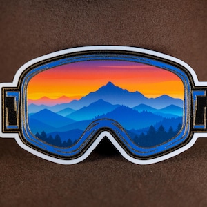 May include: A sticker featuring a stylized image of ski goggles with a mountain landscape scene inside. The scene depicts a sunset with orange and red hues, blue mountains, and a forest. The goggles have a white border with blue and gold accents.