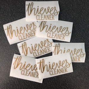 May include: A set of six white labels with the text "thieves cleaner" written in gold cursive lettering.