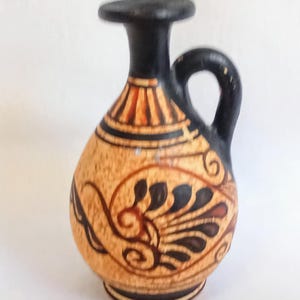 Può includere: Un piccolo vaso decorativo in ceramica con manico e beccuccio neri. Il vaso ha una base beige con dettagli dipinti in nero e rosso, tra cui un motivo floreale. La parte superiore del vaso è nera.