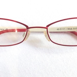 Op de afbeelding: Rode ovale bril met een dun metalen montuur. De pootjes en het bovenste deel van het montuur zijn rood, terwijl de brug en de binnenkant van het montuur gebroken wit zijn. De merknaam "Mexx" is zichtbaar. De bril is 4,9 cm breed.
