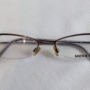 Könnte beinhalten: Eine Brille mit einem braunen Metallrahmen und klaren Gläsern. Die Brille hat ein Halbrand-Design und der Markenname "MEXX titan" ist auf dem Glas sichtbar. Die Brille liegt auf einer weißen Oberfläche.