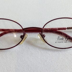 Könnte beinhalten: Ovale Brille mit einem weinroten Rahmen. Die Brille hat klare Gläser und einen dünnen Metallrahmen. Der Text "Verla Optics A TASTE FOR FINE" ist auf dem rechten Glas aufgedruckt. Sie liegt auf einer weißen Oberfläche.