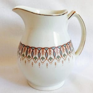 Puede incluir: Jarra de cerámica blanca con un patrón decorativo en rojo, gris y verde. La jarra tiene un borde y un asa dorados. Una pieza clásica para servir bebidas.