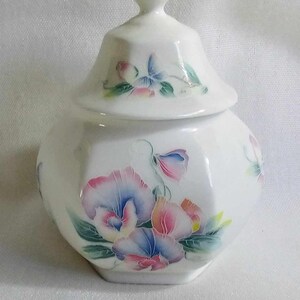 Pode incluir: Jarro de cerâmica branca com tampa, com um design floral de flores rosa, azuis e brancas. O jarro tem uma forma hexagonal.