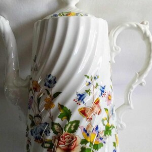 Puede incluir: Cafetera de porcelana blanca con un diseño de espiral y un patrón floral con mariposas, flores y hojas en tonos azules, amarillos, rojos y verdes.