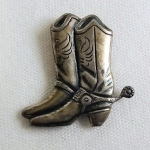 May include: A silver-toned metal pin in the shape of a pair of cowboy boots. The boots have decorative wing designs and spurs. The pin is likely a vintage or Western-themed accessory.
