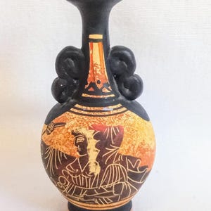 Può includere: Un piccolo vaso in ceramica nero e arancione con una dettagliata illustrazione di figure in stile classico. Il vaso ha un corpo arrotondato, un collo stretto e due manici curvi. Il design presenta una calda tavolozza di colori.