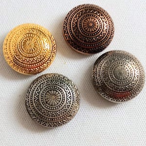 Peut inclure: Quatre boutons décoratifs circulaires. L'un est doré, l'autre est bronze et deux sont argentés. Chaque bouton présente un motif en relief détaillé de cercles concentriques et de formes géométriques.