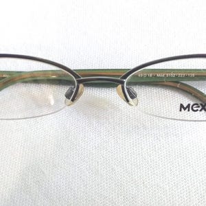 Könnte beinhalten: Halbrandlose Brille mit einem dunkelvioletten oberen Rahmen und Bügeln. Der untere Teil der Gläser ist klar. Der Markenname "MEXX" ist auf dem rechten Glas aufgedruckt. Der Rahmen hat ein grünes und goldenes Farbschema.