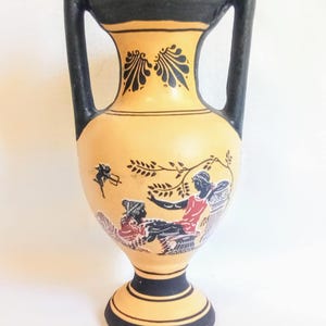 Può includere: Un vaso vintage in ceramica color crema con manici e dettagli neri. Il vaso presenta una scena dipinta con figure in rosso, nero e bianco. Il vaso ha un corpo rotondo e una base svasata, con un'altezza di circa 20 cm.