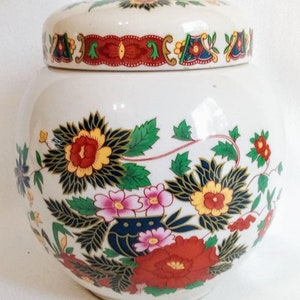 Può includere: Un vaso di ceramica bianco con coperchio, decorato con motivi floreali colorati. Il vaso ha una forma rotonda con un coperchio. Il design floreale comprende fiori rossi, gialli, blu e rosa con foglie verdi.