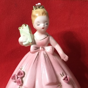 Può includere: Una statuetta in porcellana di una ragazza con un vestito rosa che tiene una scatola regalo verde. Ha i capelli biondi e una corona dorata. L'abito ha un fiocco dorato e bordi dorati. Ci sono rose rosa sull'abito.