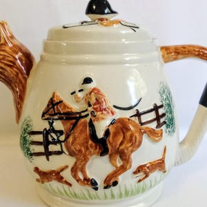 Può includere: Teiera in ceramica color crema con beccuccio a forma di testa di volpe e manico con dettaglio nero. La teiera presenta una scena di caccia con un cavaliere a cavallo, cani e una staccionata. Il coperchio ha un pomello nero.