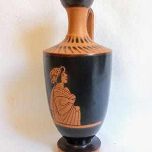 Può includere: Un vaso in ceramica nera e terracotta con manico. Il vaso presenta un design greco classico con una figura di profilo. Il vaso è alto circa 20 cm.