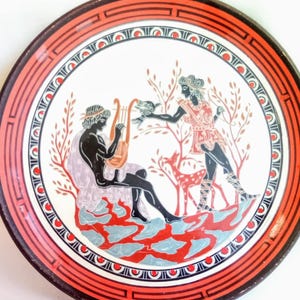 Può includere: Piatto decorativo rotondo con una scena greca classica. Il piatto presenta un bordo rosso e nero, un centro bianco con figure in nero, rosso e blu e un musicista che suona una lira. È raffigurato anche un cervo.