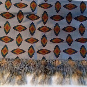 May include: A light gray rectangular scarf featuring a repeating pattern of diamond-shaped motifs in shades of orange, yellow, and dark blue. The scarf is finished with a long, textured fringe along the bottom edge in matching gray and tan tones.
