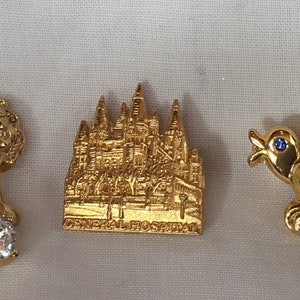 May include: Three gold-toned pins with blue accents. The first pin features a lion with a large clear rhinestone. The second pin features a building with the text "General Hospital". The third pin features a bird with a blue rhinestone eye.