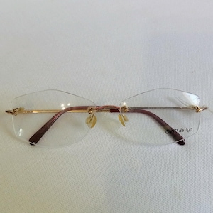 Könnte beinhalten: Randlose Brille mit goldfarbenem Steg und Bügeln sowie braunen Bügeln. Die Gläser sind klar, und die Brille hat ein angebrachtes Etikett. Der Text "OWP design" ist auf dem Bügel aufgedruckt.