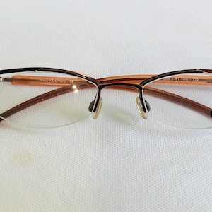 May include: Semi-rimless eyeglasses with a brown metal frame and clear lenses. The glasses have a rectangular shape with the brand name "mexx" printed on the right lens. The frame has a brown and orange color scheme.