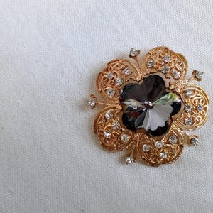 Op de afbeelding: Een broche van goudkleurige metaal met een groot, donkergrijs kristal in het midden. Het kristal is omgeven door een delicaat, versierd ontwerp van kleinere kristallen.