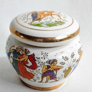 Può includere: Una scatola portaoggetti in ceramica bianca con un bordo dorato e un coperchio con una scena dipinta di persone e un cavallo. La scatola è decorata con un motivo floreale colorato.