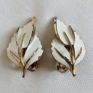 Może przedstawiać: Para klipsów z motywem liścia. Każdy kolczyk ma biały emaliowany liść ze złotymi żyłkami i obramowaniem. Kolczyki są umieszczone na białym tle.