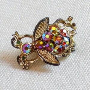 Könnte beinhalten: Eine goldfarbene Brosche mit einem fantasievollen Design, das eine stilisierte Kreatur mit schillernden Strasssteinen zeigt. Die Kreatur hat einen runden Körper und Flügel, mit grünen Akzenten an Kopf und Beinen.