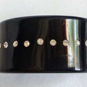 Peut inclure: Un large bracelet noir orné d'une rangée de petits strass étincelants. Le bracelet a une forme cylindrique solide, et les strass sont espacés uniformément autour du bracelet. La surface du bracelet est brillante.