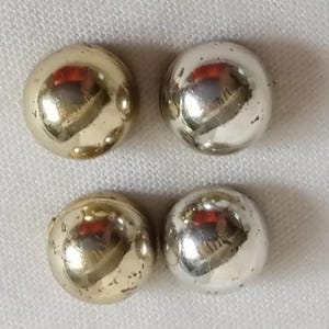 以下が含まれることがあります： 4つの小さくて丸い金属製のビーズ。2つは金色で、2つは銀色です。ビーズは、白くテクスチャ加工された表面に正方形のパターンで配置されています。ビーズは周囲の環境を反射しています。