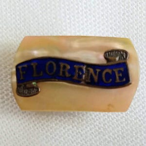 May include: A rectangular pin with the word "FLORENCE" on a blue banner. The pin has a pearlescent background and decorative metal accents.
