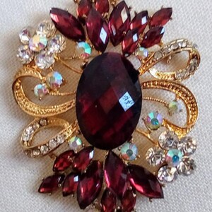 May include: A gold-toned brooch with a large, faceted, dark red gemstone surrounded by smaller red and clear rhinestones. The brooch has a floral design with intricate details.