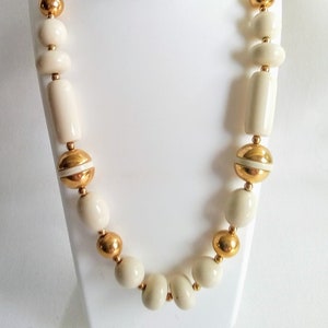 May include: A white and gold beaded necklace with round and cylindrical beads. The necklace is on a white mannequin.
