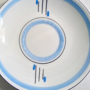 May include: A white saucer with a blue and black design. The saucer has a blue band around the edge and a blue circle in the center with a black line design.