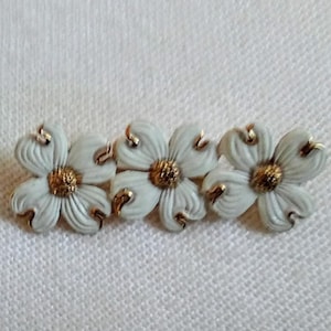 May include: A white porcelain brooch featuring three gold-trimmed flowers. The flowers are arranged in a row and have a delicate, feminine design.