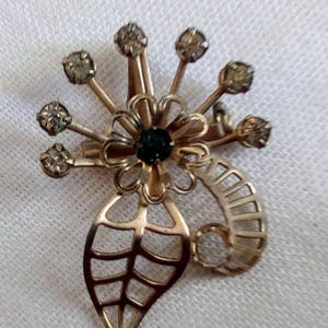 May include: A gold-toned brooch with a green gem in the center, surrounded by a flower design and a leaf with a geometric pattern. The brooch is adorned with clear rhinestones.
