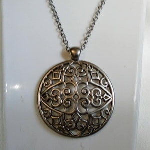 Op de afbeelding: Een zilveren ketting met een grote, ronde hanger met een delicaat, ingewikkeld filigraan ontwerp.