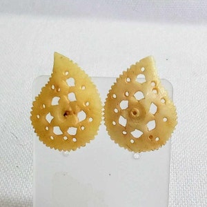May include: A pair of cream-colored earrings made of carved shell. The earrings are shaped like teardrops with intricate cut-out designs.