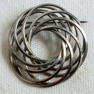 May include: A silver brooch featuring a circular design made from interwoven, curved metal bands. The brooch has a pin fastening. The design resembles a stylized flower or spiral.