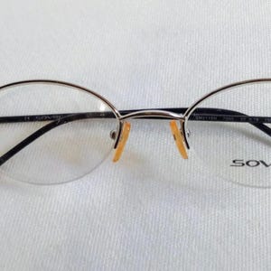 May include: Oval-shaped eyeglasses with a silver-colored frame and black temples. The lenses are clear, and the brand name "sover" is printed on the right lens. The glasses are set against a white background.