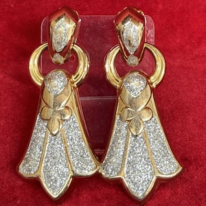 Könnte beinhalten: Ein Paar goldfarbene Ohrringe mit einem funkelnden, kristallartigen Design. Die Ohrringe haben eine geometrische Form mit einem blütenartigen Detail und einem baumelnden, glockenförmigen Boden. Die Ohrringe sind vor rotem Hintergrund platziert.