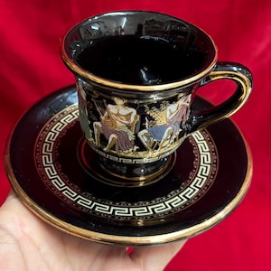 Op de afbeelding: Zwarte keramische theekop en schotelset met gouden rand en Griekse figuren. De kop en schotel hebben een zwarte achtergrond met gouden details en een zich herhalend geometrisch patroon rond de rand.